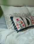 Garden Blossom Cushion Cover – This handcrafted cotton cushion cover features intricate floral embroidery inspired by traditional Mughal motifs. The delicate pink and orange blossoms contrast elegantly against a soft cream background, framed with deep blue detailing and finished with playful tassel accents.