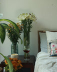 A cozy bedside setup featuring fresh white lilies and yellow wildflowers in tall glass vases, adding a touch of nature to the space. A textured glass of red wine sits on a dark wooden side table, alongside a ceramic bowl filled with snacks. Soft linen bedding with delicate hand embroidery completes the tranquil and inviting ambiance.