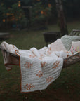 A cozy outdoor setup featuring the Amber Bloom Quilt, delicately draped over a hammock. Handwoven from soft cotton and adorned with intricate hand-block printed floral motifs in warm amber hues, this quilt adds a touch of artisanal charm to a tranquil garden retreat. Paired with embroidered pillows, it creates an inviting space to relax and unwind.