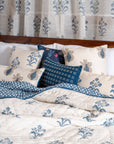 A serene bedroom setting featuring the Indigo Reverie Quilt, handwoven in cotton and adorned with delicate hand block-printed floral motifs in a soothing indigo hue. Layered with coordinating pillows and curtains, this arrangement exudes timeless elegance, bringing a touch of artisanal craftsmanship and refined comfort to the space.