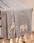 The Serene Elephant Throw, a beautifully handwoven textile adorned with delicate hand-embroidered elephant motifs. Its soft grey fabric is complemented by intricate detailing and oversized ivory tassels, adding a touch of warmth and artisanal charm to any space.