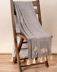 The Serene Elephant Throw, a beautifully handwoven textile adorned with delicate hand-embroidered elephant motifs. Its soft grey fabric is complemented by intricate detailing and oversized ivory tassels, adding a touch of warmth and artisanal charm to any space.