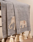 The Serene Elephant Throw, a beautifully handwoven textile adorned with delicate hand-embroidered elephant motifs. Its soft grey fabric is complemented by intricate detailing and oversized ivory tassels, adding a touch of warmth and artisanal charm to any space.