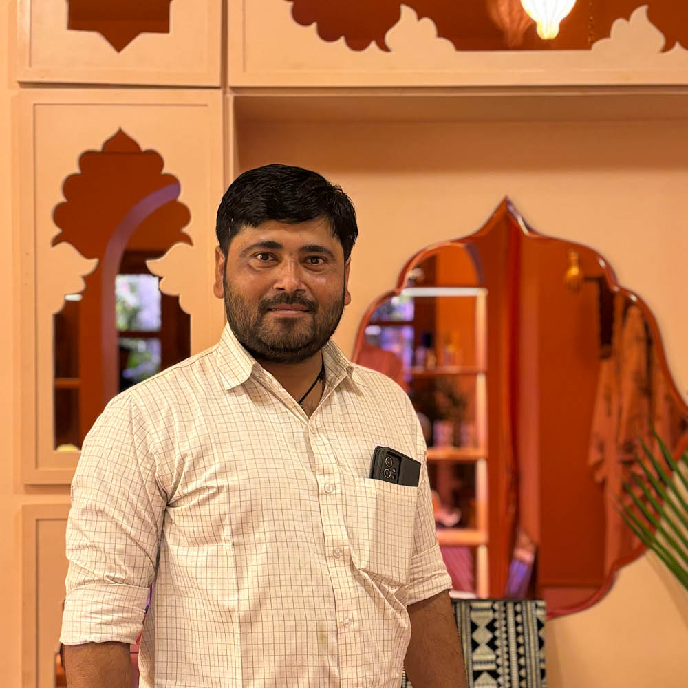 A skilled artisan standing in front of a beautifully designed space with traditional Indian arch motifs. Dressed in a checkered shirt with a phone tucked into his pocket, he carries a warm and confident presence, reflecting the craftsmanship and heritage he represents. The rich, warm tones of the background mirror the artistry woven into his work.