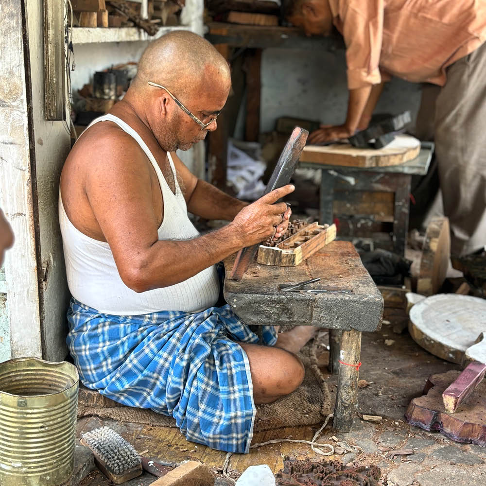 A master artisan meticulously carving a wooden block by hand, shaping intricate patterns for traditional block printing. Seated on a low wooden stool, he works with precision, surrounded by tools and raw materials in his workshop. The scene reflects the dedication and craftsmanship passed down through generations, where each block becomes a piece of art in itself.