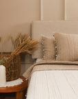 A serene bedroom setting featuring the Kantha Textured Stories Bedspread, delicately hand-stitched with multicolored threads on a soft, textured surface. The bedding is complemented by matching pillows adorned with handcrafted pom-poms, adding warmth and character. A woven basket filled with dried wheat and a rustic wooden side table enhance the cozy, natural ambiance.