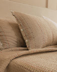 A cozy setting featuring the Kantha Textured Stories Bedspread, adorned with delicate hand-stitched details in multicolored threads. The soft, textured fabric pairs beautifully with matching pillows, finished with playful handcrafted pom-pom accents, creating a warm and inviting atmosphere.