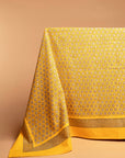 The Block-Printed Yellow Tablecloth drapes elegantly over a table, its intricate geometric pattern radiating warmth and craftsmanship. Framed with a bold yellow border, this piece brings a touch of artisanal charm to any dining space.