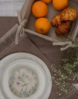 A serene breakfast setting featuring a handwoven linen table runner and matching table mats with delicate frayed edges. A basket filled with fresh oranges and flaky croissants sits atop, while a white ceramic bowl with a floral-painted interior adds an artistic touch. Sprigs of baby's breath complete the scene, blending nature with timeless craftsmanship.