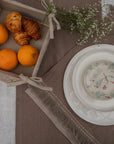 A serene breakfast setting featuring a handwoven linen table runner and matching table mats with delicate frayed edges. A basket filled with fresh oranges and flaky croissants sits atop, while a white ceramic bowl with a floral-painted interior adds an artistic touch. Sprigs of baby's breath complete the scene, blending nature with timeless craftsmanship.