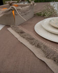 A serene breakfast setting featuring a handwoven linen table runner and matching table mats with delicate frayed edges. A basket filled with fresh oranges and flaky croissants sits atop, while a white ceramic bowl with a floral-painted interior adds an artistic touch. Sprigs of baby's breath complete the scene, blending nature with timeless craftsmanship.