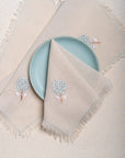 The Veiled Flower Napkin exudes understated elegance with its soft beige cotton weave and delicate embroidery. A single hand-stitched flower in muted blue and white graces the fabric, adding a refined touch to any dining setting. Thoughtfully crafted, its texture and minimalist design celebrate timeless sophistication.
