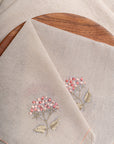 The Misty Garden Napkin brings delicate charm to the dining table with its soft, airy weave and intricate floral embroidery. Hand-stitched blossoms in warm pink tones, complemented by muted green leaves, add a subtle yet elegant touch. Finished with fine fringed edges, this napkin embodies effortless sophistication.