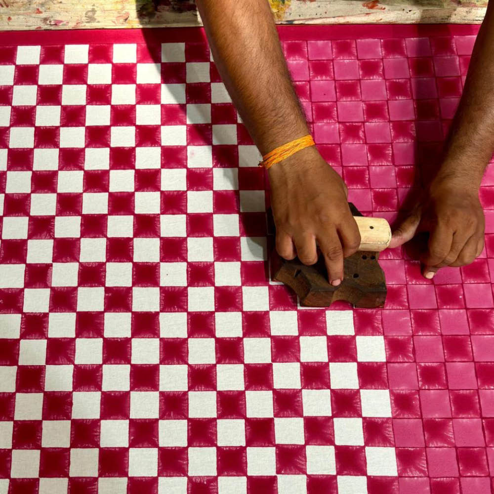 An artisan carefully hand-block prints a bold red and white checkerboard pattern onto fabric using a carved wooden block. The rhythmic stamping process brings the design to life, with each press leaving behind a perfectly aligned imprint. The precision and patience of traditional block printing shine through in this vibrant, handcrafted textile.