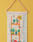 Climber Height Chart – A beautifully handcrafted cotton height chart featuring a vibrant illustration of a wooden plant shelf adorned with lush greenery, colorful pots, and playful garden elements like a straw hat, dragonfly, and butterfly. Hand-stitched details add texture and charm, making it a delightful keepsake for tracking growth milestones.