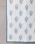 The Indigo Reverie Quilt, crafted from handwoven cotton, showcases a delicate floral pattern meticulously hand block printed in a serene indigo hue. Its soft, quilted texture and refined border add a timeless elegance, making it a perfect addition to any tranquil and inviting space.