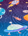 A close-up of the "Tim, the Traveler Quilt," a handcrafted space-themed quilt featuring an appliqué design of a vibrant blue and green planet, a fiery comet, and scattered stars on a deep purple background. The quilt is folded to reveal its playful geometric patterned reverse side in shades of blue, purple, and orange. Placed on a soft white textured surface, an astronaut figurine lies beside it, enhancing the celestial theme.