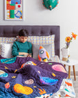 A cozy and imaginative bedroom scene featuring a young boy reading under the "Tim, the Traveler Quilt." The quilt, handcrafted with intricate space-themed appliqué, showcases planets, rockets, and stars in vibrant colors. A matching rocket-shaped cushion adds to the celestial theme, while plaid pillows and a dark upholstered headboard provide a modern contrast. A side table with fresh flowers, a quirky astronaut figurine, and a framed sports-inspired artwork complete the playful yet stylish setting.