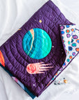 A close-up of the "Tim, the Traveler Quilt," a handcrafted space-themed quilt featuring an appliqué design of a vibrant blue and green planet, a fiery comet, and scattered stars on a deep purple background. The quilt is folded to reveal its playful geometric patterned reverse side in shades of blue, purple, and orange. Placed on a soft white textured surface, an astronaut figurine lies beside it, enhancing the celestial theme.