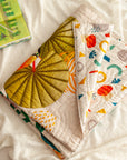 A folded The Forest is My Playground quilt, handcrafted from soft cotton, featuring a vibrant jungle scene with a toucan perched under large tropical leaves. The quilt’s reverse side displays a playful abstract pattern in bright, modern colors. Set against a cozy white blanket with a children's book nearby, the image captures warmth, comfort, and storytelling through textile artistry.