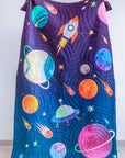 A cozy and imaginative bedroom scene featuring a young boy reading under the "Tim, the Traveler Quilt." The quilt, handcrafted with intricate space-themed appliqué, showcases planets, rockets, and stars in vibrant colors. A matching rocket-shaped cushion adds to the celestial theme, while plaid pillows and a dark upholstered headboard provide a modern contrast. A side table with fresh flowers, a quirky astronaut figurine, and a framed sports-inspired artwork complete the playful yet stylish setting.