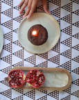 A cozy table setting featuring the Black Triangles Tablecloth, a hand-block printed cotton piece with a striking geometric pattern. The warm, earthy tones provide a perfect backdrop for a chocolate-covered dessert with a lit candle, a wooden serving tray with a freshly cut pomegranate, and ceramic plates. A moment of indulgence, beautifully framed by artisanal craftsmanship.