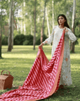 A woman stands in a sunlit grove, gracefully holding the Pink Squares Tablecloth, its hand-dyed hues catching the light. The picnic scene unfolds beside her, with a woven basket resting on the grass, inviting a moment of leisure and warmth amidst nature.