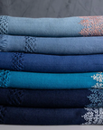 A neatly stacked selection of Chambi Bedspreads with Snow Shades, each in varying shades of blue, adorned with intricate embroidery and delicate fringe details. The gradient of hues, from sky blue to deep navy, showcases the fine craftsmanship and timeless elegance of these handcrafted textiles.