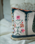 Garden Blossom Cushion Cover – This handcrafted cotton cushion cover features intricate floral embroidery inspired by traditional Mughal motifs. The delicate pink and orange blossoms contrast elegantly against a soft cream background, framed with deep blue detailing and finished with playful tassel accents.