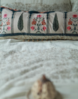 Garden Blossom Cushion Cover – This handcrafted cotton cushion cover features intricate floral embroidery inspired by traditional Mughal motifs. The delicate pink and orange blossoms contrast elegantly against a soft cream background, framed with deep blue detailing and finished with playful tassel accents.