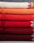 A stack of Chambi Bedspreads, featuring a rich gradient of warm hues, from ivory and burnt orange to deep crimson and brown. Each piece showcases intricate embroidery along the edges, complemented by delicate fringe details. Among them is the Chambi Bedspread in Brown and White, embodying timeless elegance and artisanal craftsmanship.