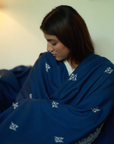 A woman wrapped in the Chambi Bedspread with Snow Shades, a deep blue textile adorned with delicate white embroidery. The soft fabric drapes around her, evoking warmth and comfort in a serene indoor setting.