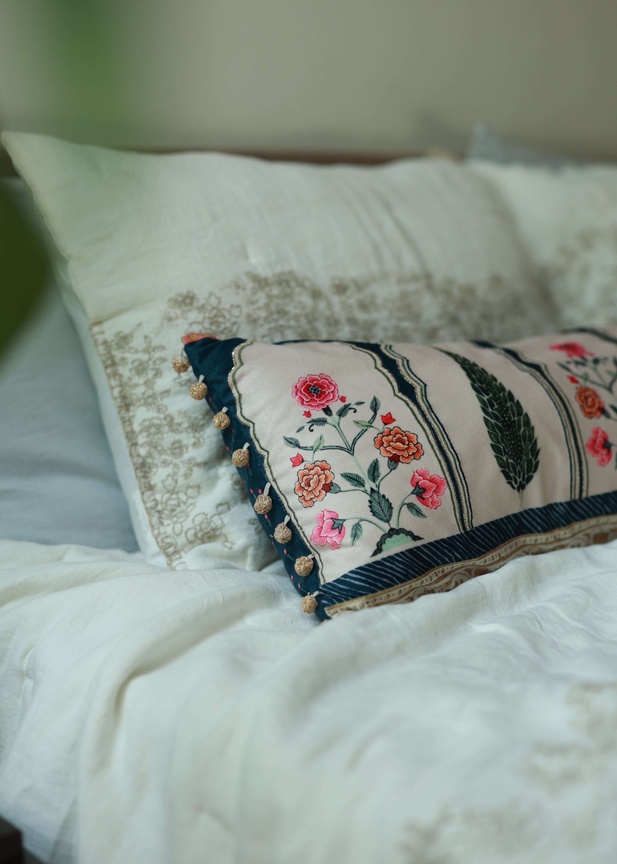 Garden Blossom Cushion Cover – This handcrafted cotton cushion cover features intricate floral embroidery inspired by traditional Mughal motifs. The delicate pink and orange blossoms contrast elegantly against a soft cream background, framed with deep blue detailing and finished with playful tassel accents.
