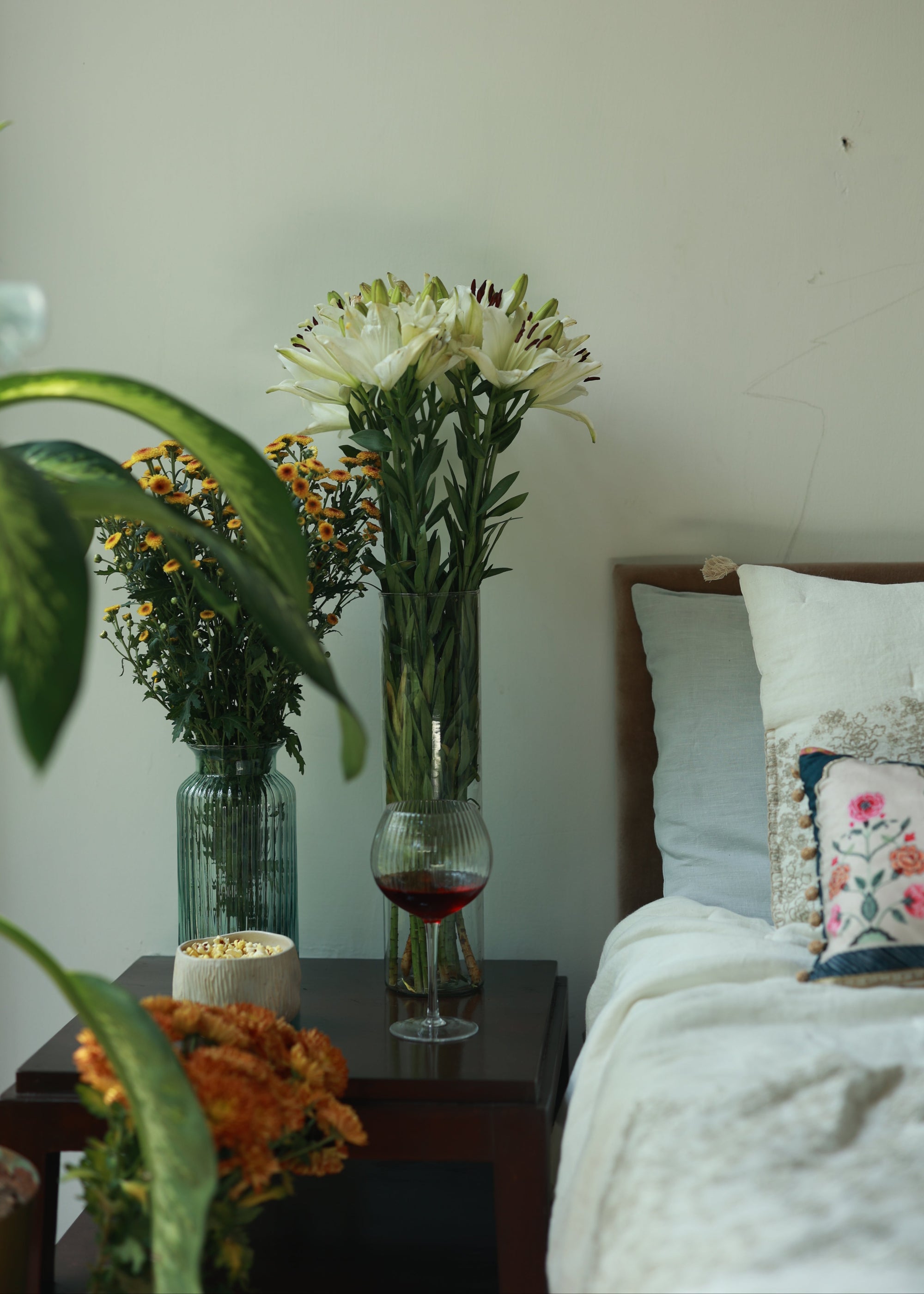 A cozy bedside setup featuring fresh white lilies and yellow wildflowers in tall glass vases, adding a touch of nature to the space. A textured glass of red wine sits on a dark wooden side table, alongside a ceramic bowl filled with snacks. Soft linen bedding with delicate hand embroidery completes the tranquil and inviting ambiance.