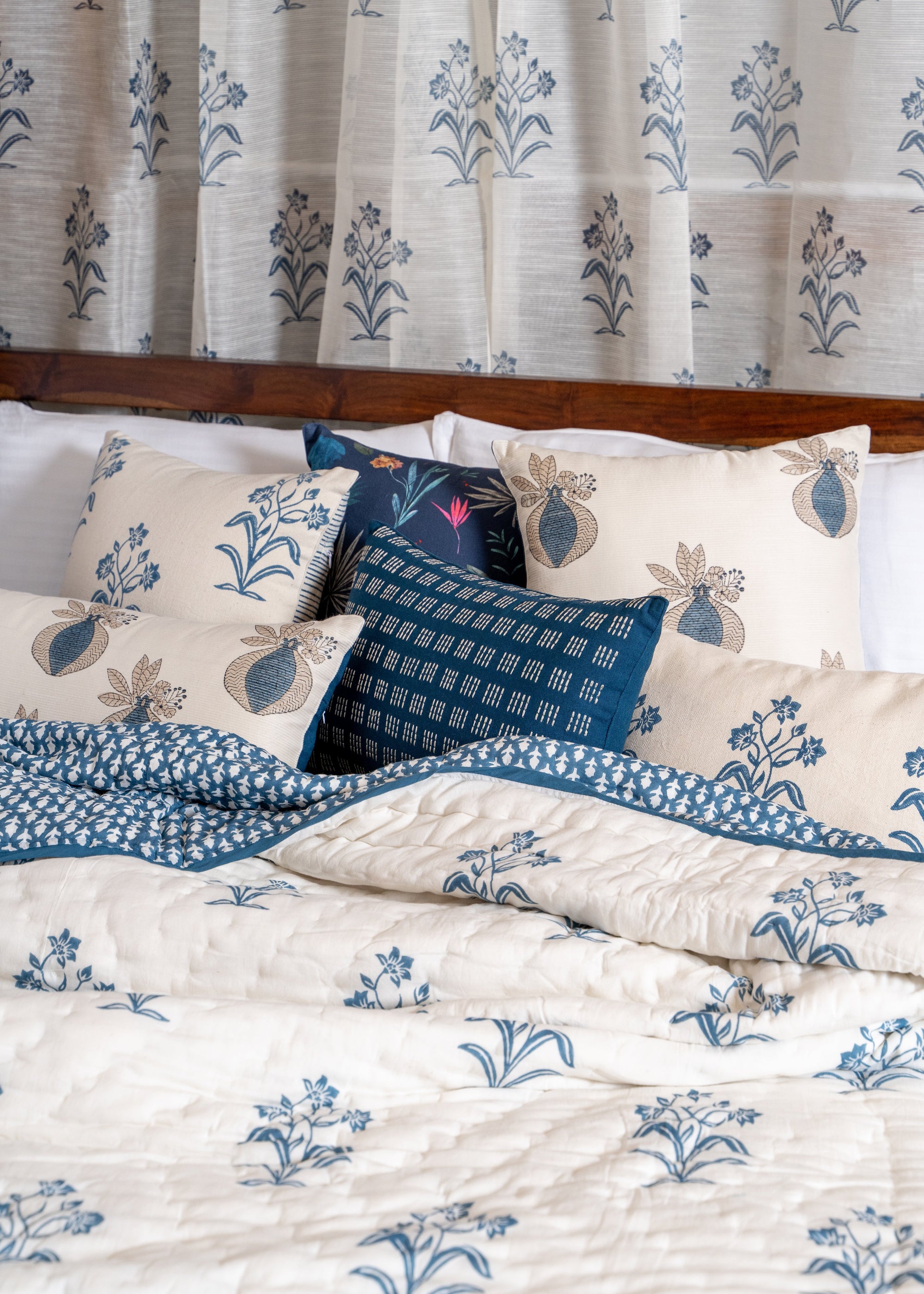 A serene bedroom setting featuring the Indigo Reverie Quilt, handwoven in cotton and adorned with delicate hand block-printed floral motifs in a soothing indigo hue. Layered with coordinating pillows and curtains, this arrangement exudes timeless elegance, bringing a touch of artisanal craftsmanship and refined comfort to the space.