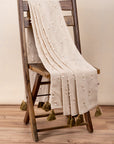 The Bowtie Embrace Throw, a handwoven cotton textile adorned with delicate hand-embroidered bowtie details. Finished with olive-green tassels, this throw brings a soft, textured charm to any space, blending artisanal craftsmanship with understated elegance.