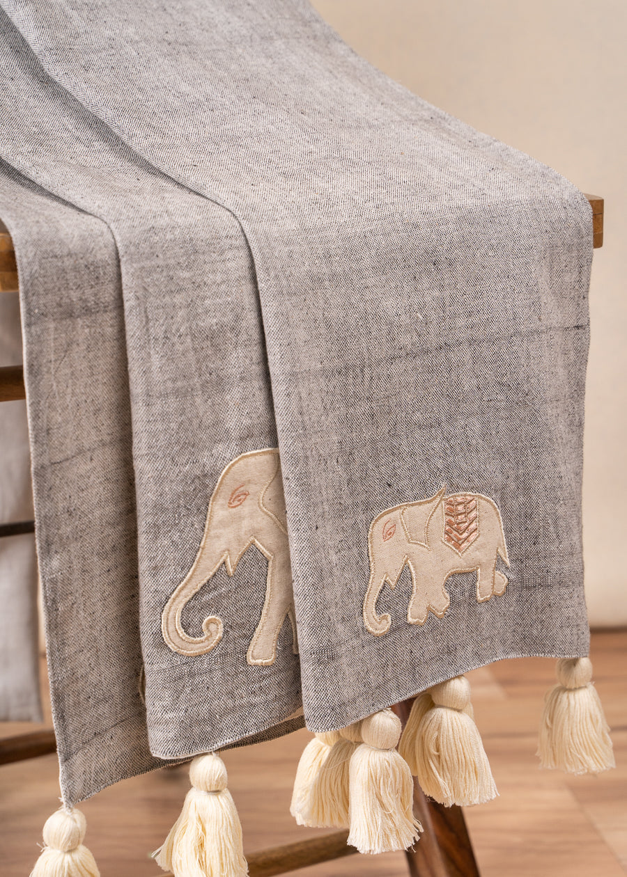 The Serene Elephant Throw, a beautifully handwoven textile adorned with delicate hand-embroidered elephant motifs. Its soft grey fabric is complemented by intricate detailing and oversized ivory tassels, adding a touch of warmth and artisanal charm to any space.