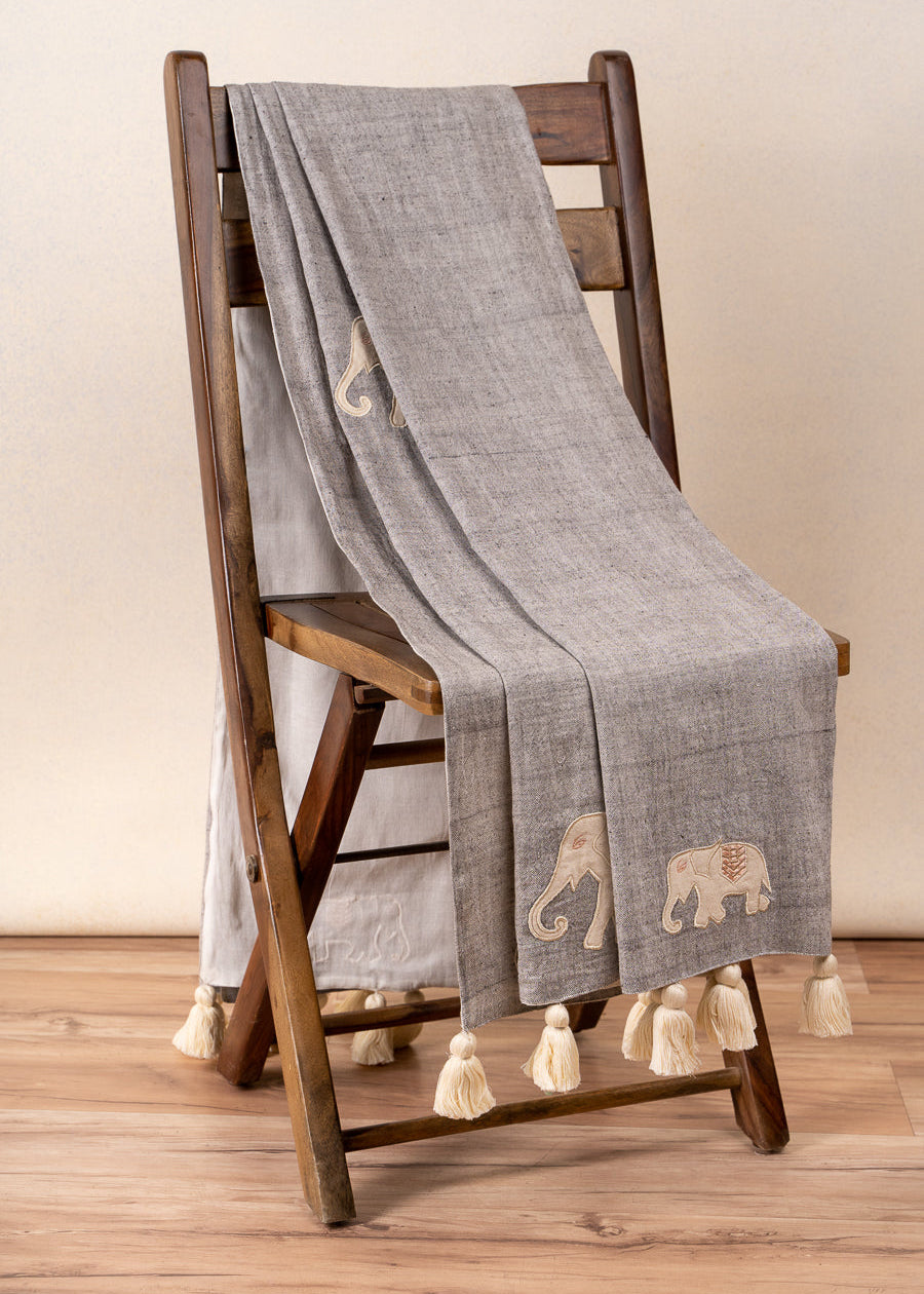 The Serene Elephant Throw, a beautifully handwoven textile adorned with delicate hand-embroidered elephant motifs. Its soft grey fabric is complemented by intricate detailing and oversized ivory tassels, adding a touch of warmth and artisanal charm to any space.