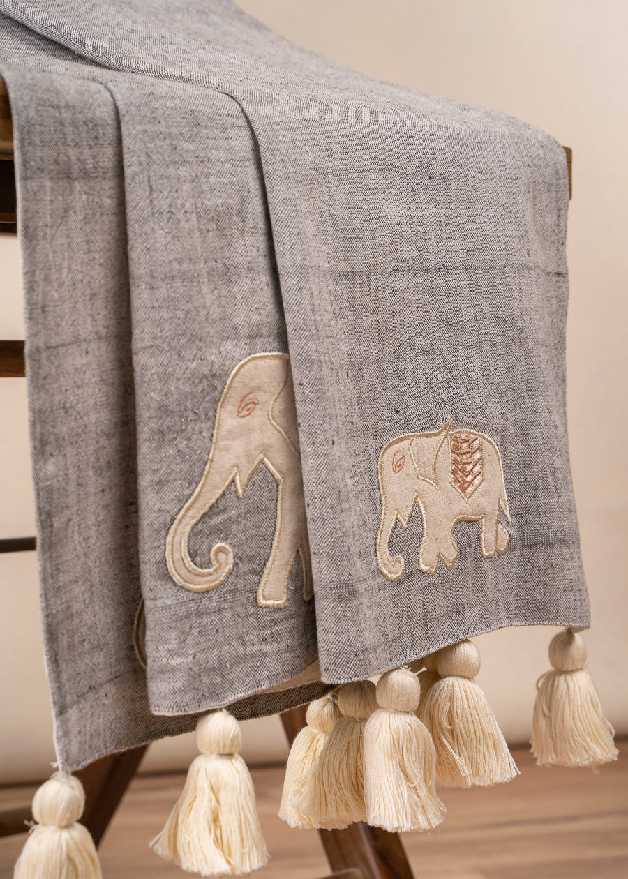 The Serene Elephant Throw, a beautifully handwoven textile adorned with delicate hand-embroidered elephant motifs. Its soft grey fabric is complemented by intricate detailing and oversized ivory tassels, adding a touch of warmth and artisanal charm to any space.
