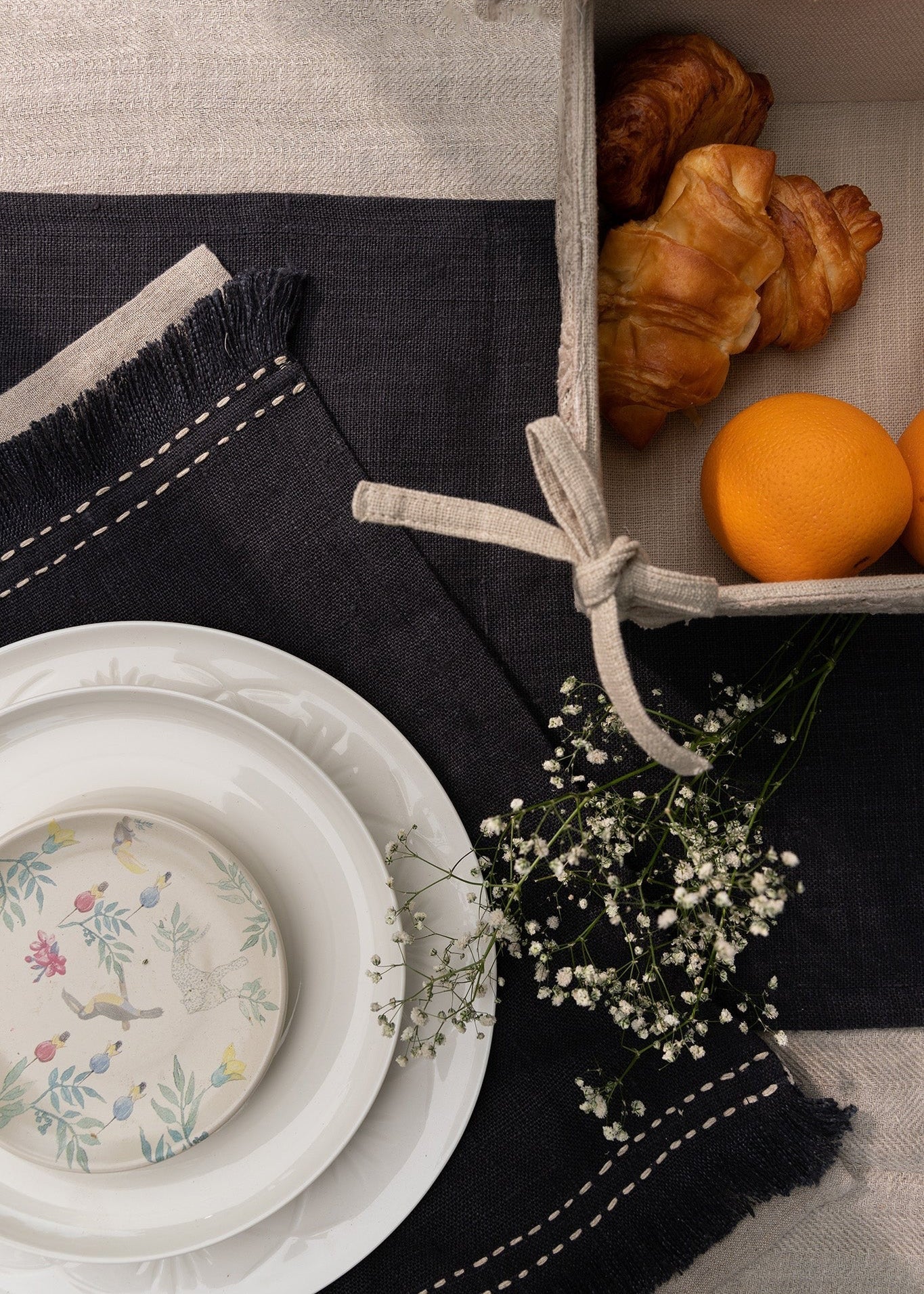 A sophisticated tablescape featuring a handwoven charcoal linen table runner and matching table mats with delicate frayed edges and hand-stitched detailing. A white ceramic plate with an intricate floral-painted interior adds a touch of softness against the deep fabric tones, blending timeless craftsmanship with modern elegance.