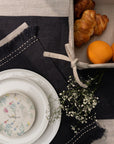 A sophisticated tablescape featuring a handwoven charcoal linen table runner and matching table mats with delicate frayed edges and hand-stitched detailing. A white ceramic plate with an intricate floral-painted interior adds a touch of softness against the deep fabric tones, blending timeless craftsmanship with modern elegance.
