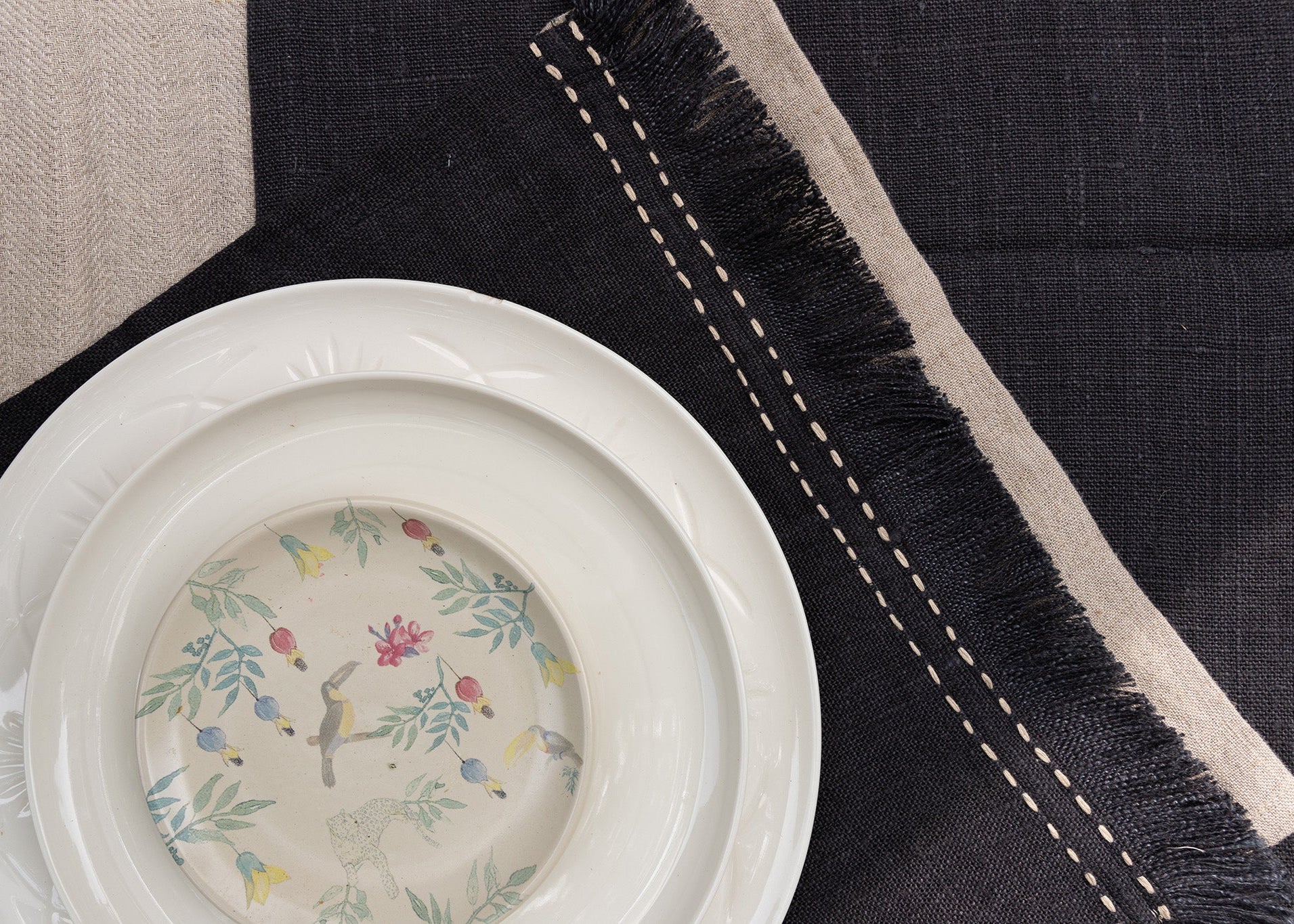 A sophisticated tablescape featuring a handwoven charcoal linen table runner and matching table mats with delicate frayed edges and hand-stitched detailing. A white ceramic plate with an intricate floral-painted interior adds a touch of softness against the deep fabric tones, blending timeless craftsmanship with modern elegance.