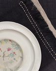A sophisticated tablescape featuring a handwoven charcoal linen table runner and matching table mats with delicate frayed edges and hand-stitched detailing. A white ceramic plate with an intricate floral-painted interior adds a touch of softness against the deep fabric tones, blending timeless craftsmanship with modern elegance.