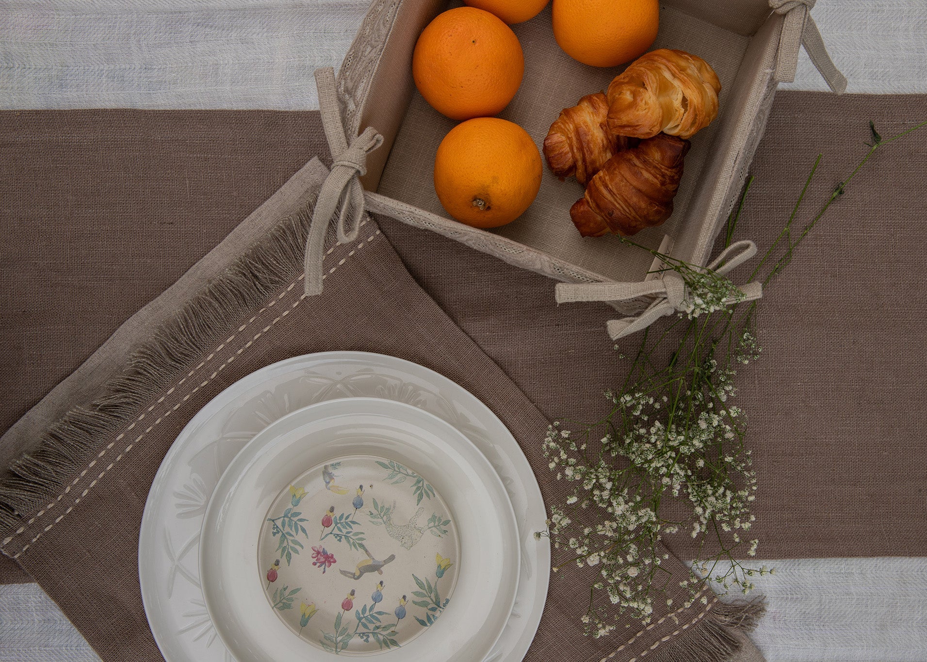 A serene breakfast setting featuring a handwoven linen table runner and matching table mats with delicate frayed edges. A basket filled with fresh oranges and flaky croissants sits atop, while a white ceramic bowl with a floral-painted interior adds an artistic touch. Sprigs of baby's breath complete the scene, blending nature with timeless craftsmanship.
