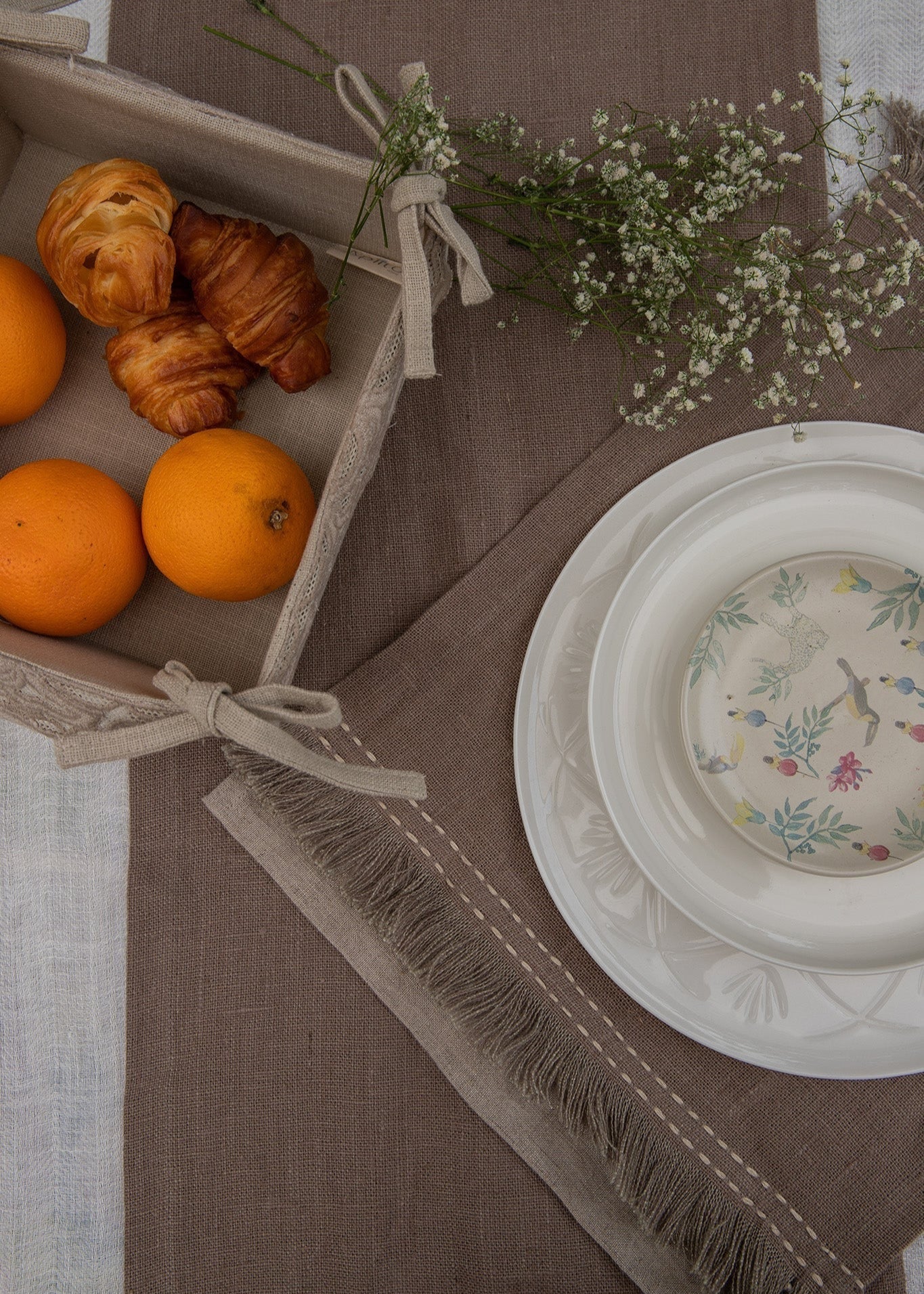 A serene breakfast setting featuring a handwoven linen table runner and matching table mats with delicate frayed edges. A basket filled with fresh oranges and flaky croissants sits atop, while a white ceramic bowl with a floral-painted interior adds an artistic touch. Sprigs of baby's breath complete the scene, blending nature with timeless craftsmanship.