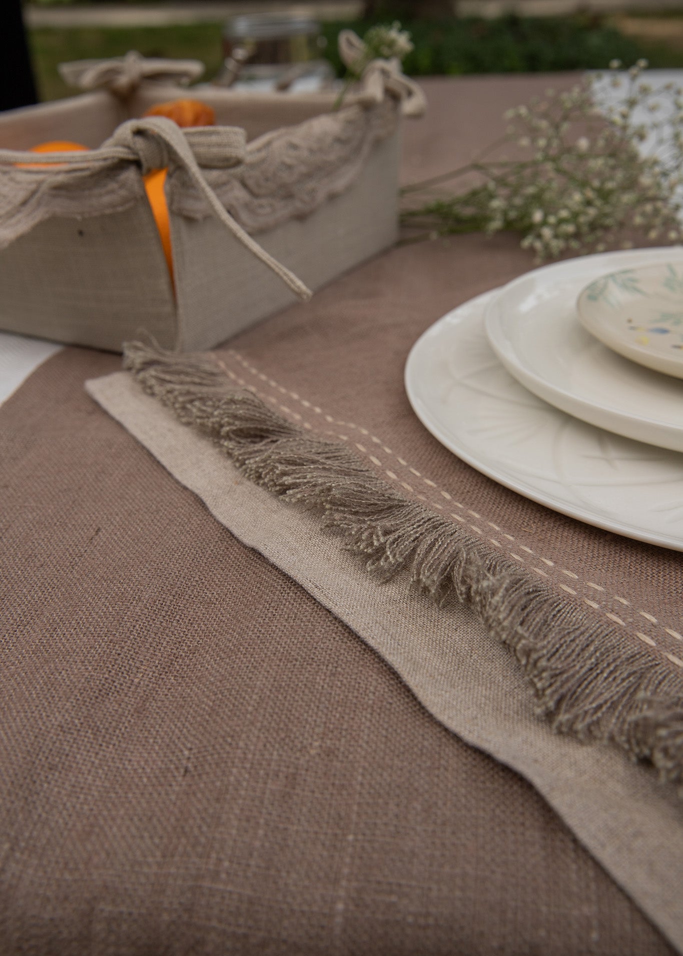 A serene breakfast setting featuring a handwoven linen table runner and matching table mats with delicate frayed edges. A basket filled with fresh oranges and flaky croissants sits atop, while a white ceramic bowl with a floral-painted interior adds an artistic touch. Sprigs of baby's breath complete the scene, blending nature with timeless craftsmanship.