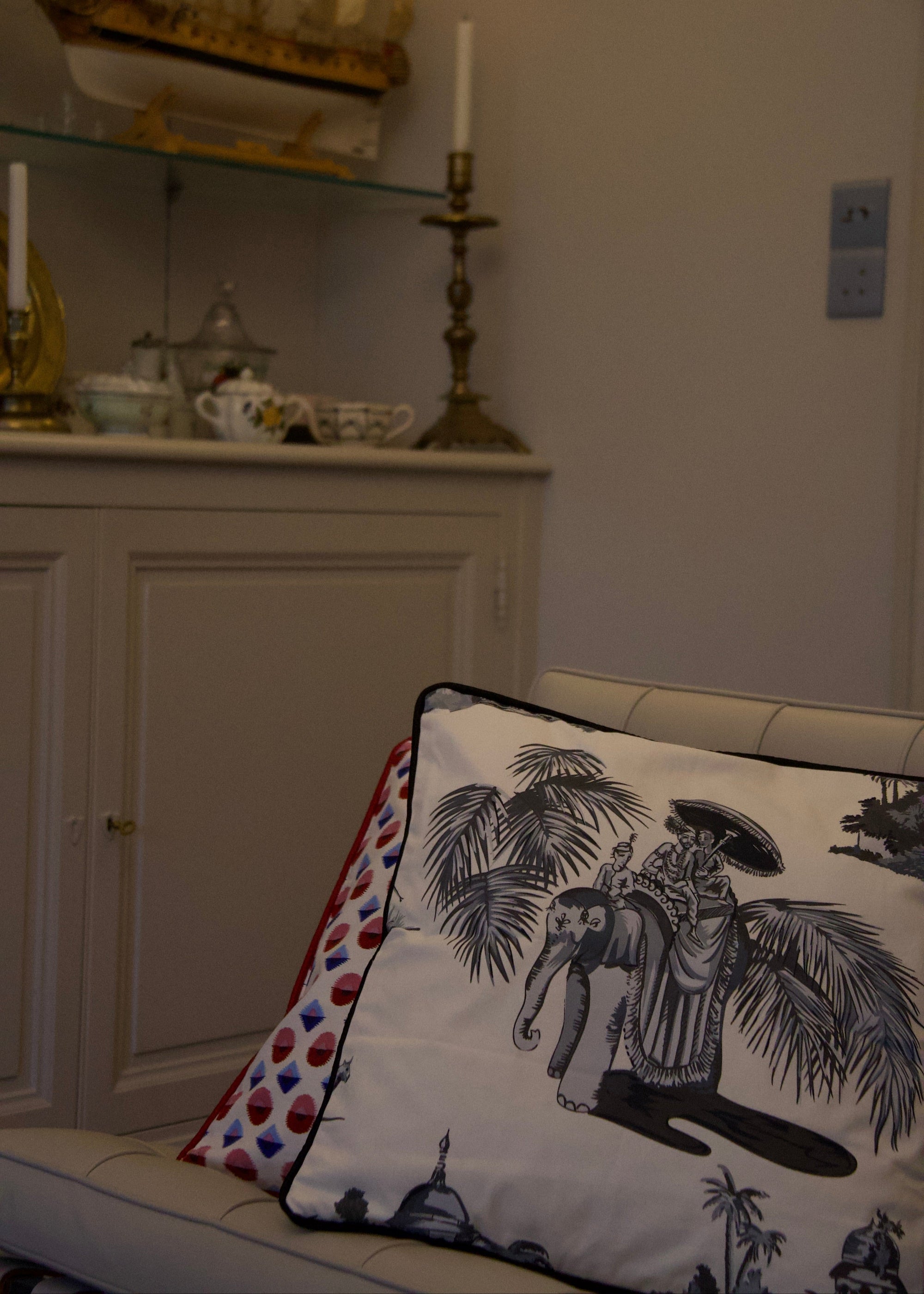 A duo of handcrafted cushions resting on a beige sofa, featuring the Heritage Threads Cushion Cover depicting a black and white Mughal elephant procession, and the Geometric Red Cushion Cover with a bold red and blue pattern. A fusion of heritage craftsmanship and contemporary design, each piece tells a unique story.