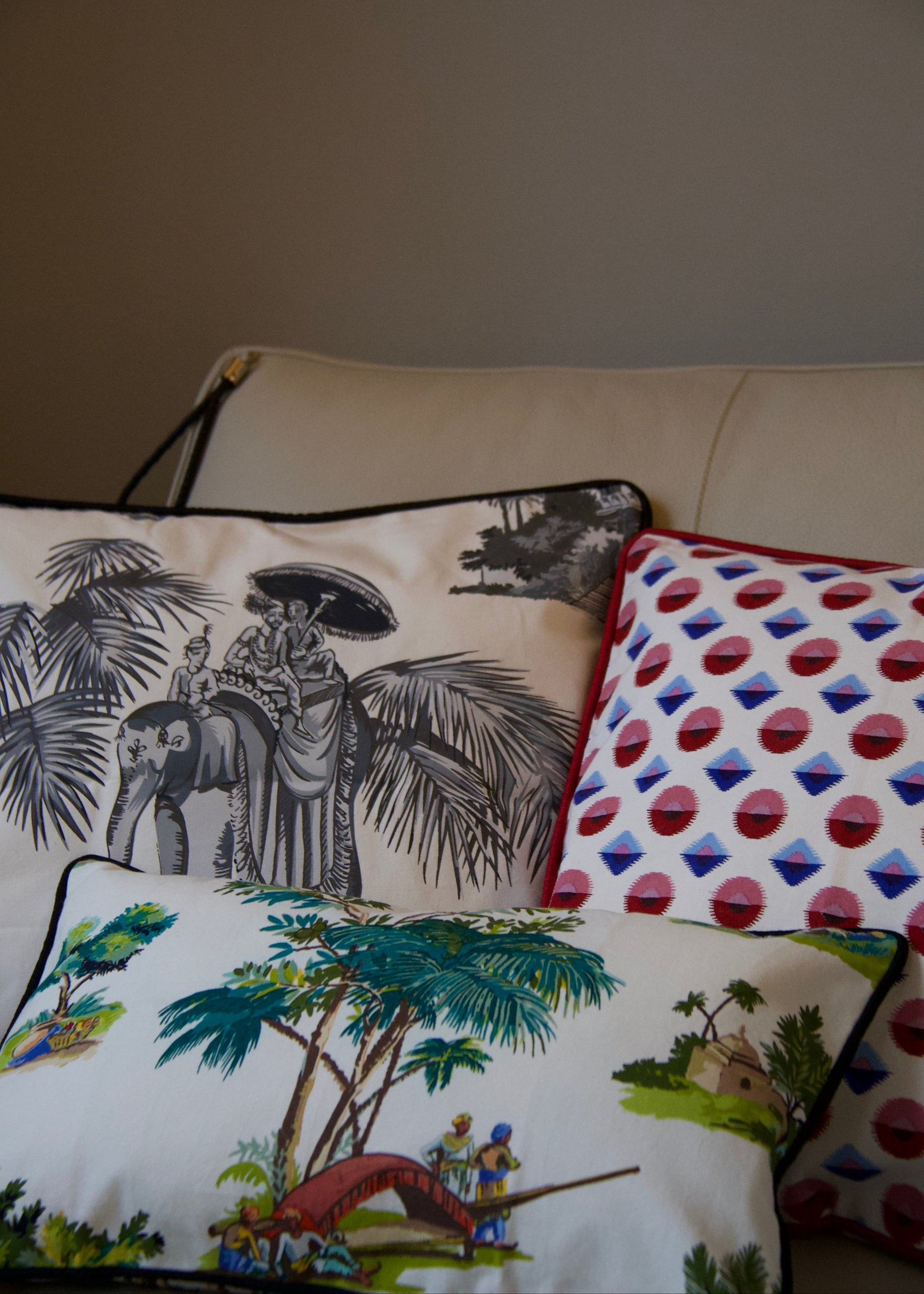 A trio of handcrafted cushions resting on a beige sofa, featuring the Mughal Orchard Cushion Cover with a lush tropical landscape, the Heritage Threads Cushion Cover depicting a black and white Mughal elephant procession, and the Geometric Red Cushion Cover with a bold red and blue pattern. A fusion of heritage craftsmanship and contemporary design, each piece tells a unique story.