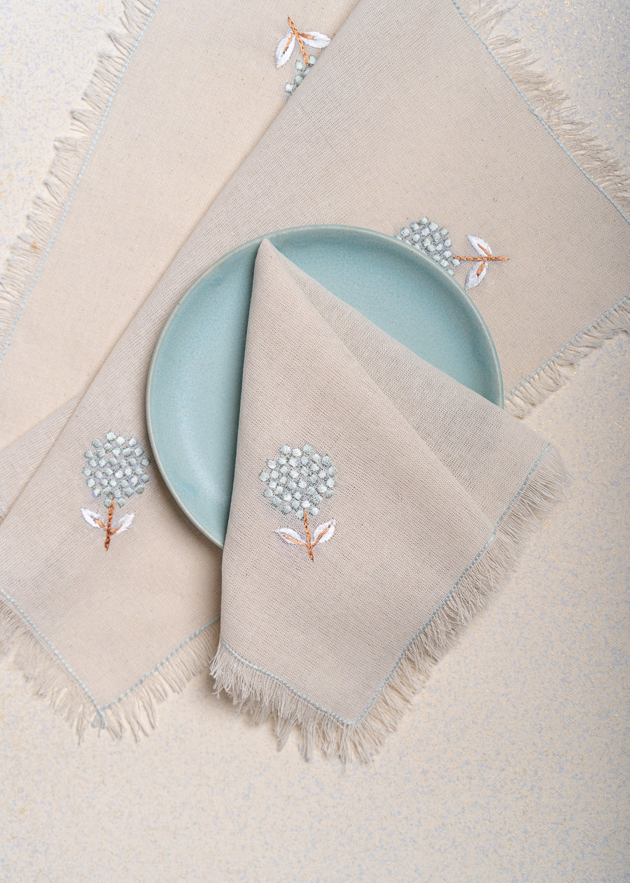 The Veiled Flower Napkin exudes understated elegance with its soft beige cotton weave and delicate embroidery. A single hand-stitched flower in muted blue and white graces the fabric, adding a refined touch to any dining setting. Thoughtfully crafted, its texture and minimalist design celebrate timeless sophistication.