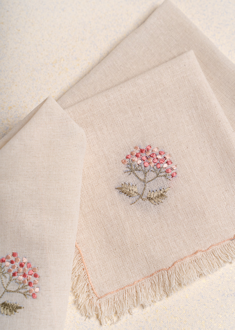 The Misty Garden Napkin brings delicate charm to the dining table with its soft, airy weave and intricate floral embroidery. Hand-stitched blossoms in warm pink tones, complemented by muted green leaves, add a subtle yet elegant touch. Finished with fine fringed edges, this napkin embodies effortless sophistication.
